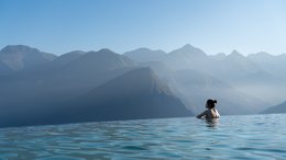 Hotel Kronblick Frau in Infinity-Pool mit Blick auf nebelige Berglandschaft bei klarem Himmel