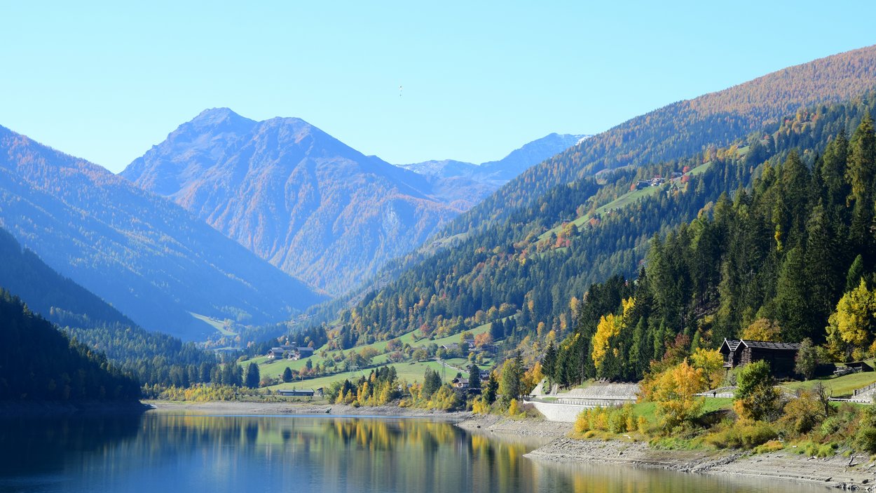Der Zoggler-See im Ultental Bergsee mit Wald und Hütten im Herbst unter klarem Himmel