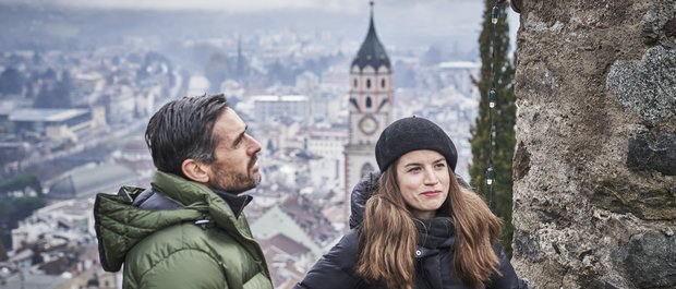 Hotel in Alto Adige 😎☀️ Per vacanze indimenticabili Coppia con abbigliamento invernale davanti a una città con torre della chiesa sullo sfondo