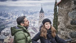 Hotels in Alta Badia: Urlaub durch und durch Paar in Winterkleidung vor einer Stadtansicht mit Kirchturm im Hintergrund