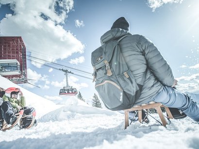 Rodeln in Südtirol: da schlägt das Herz schneller. Zwei Kinder und ein Erwachsener rodeln im Schnee bei Sonnenschein mit Seilbahn im Hintergrund