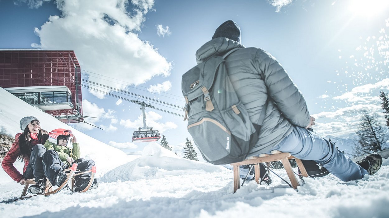 Spaß für die ganze Familie Zwei Kinder und ein Erwachsener rodeln im Schnee bei Sonnenschein mit Seilbahn im Hintergrund