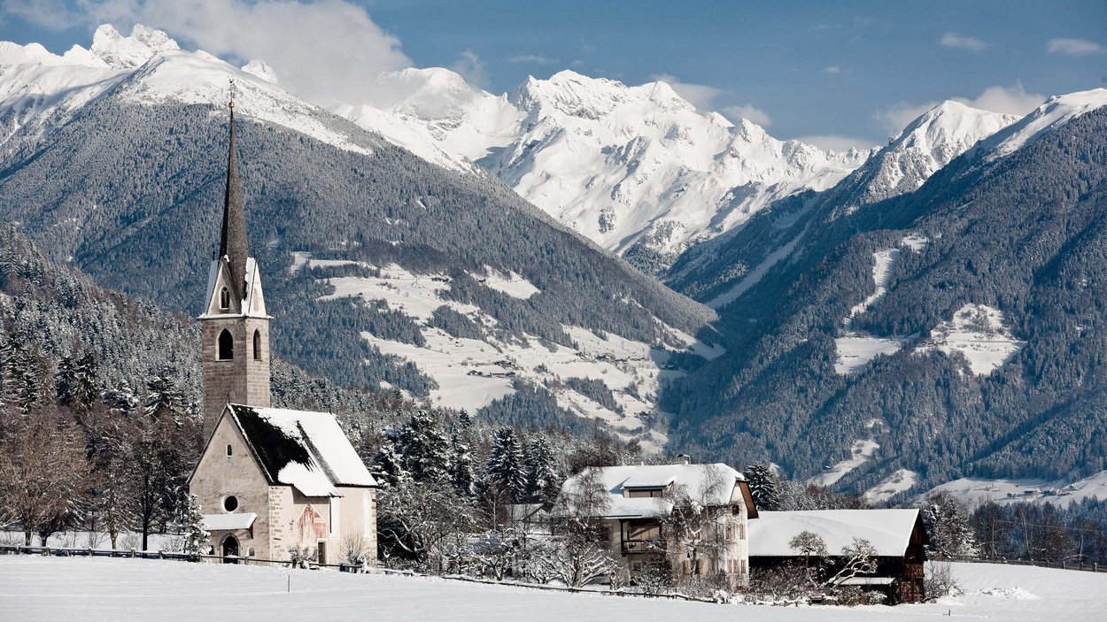 Der Kronplatz – grenzenloses Langlaufvergnügen Schneebedeckte Kirche und Häuser vor Berglandschaft im Winter