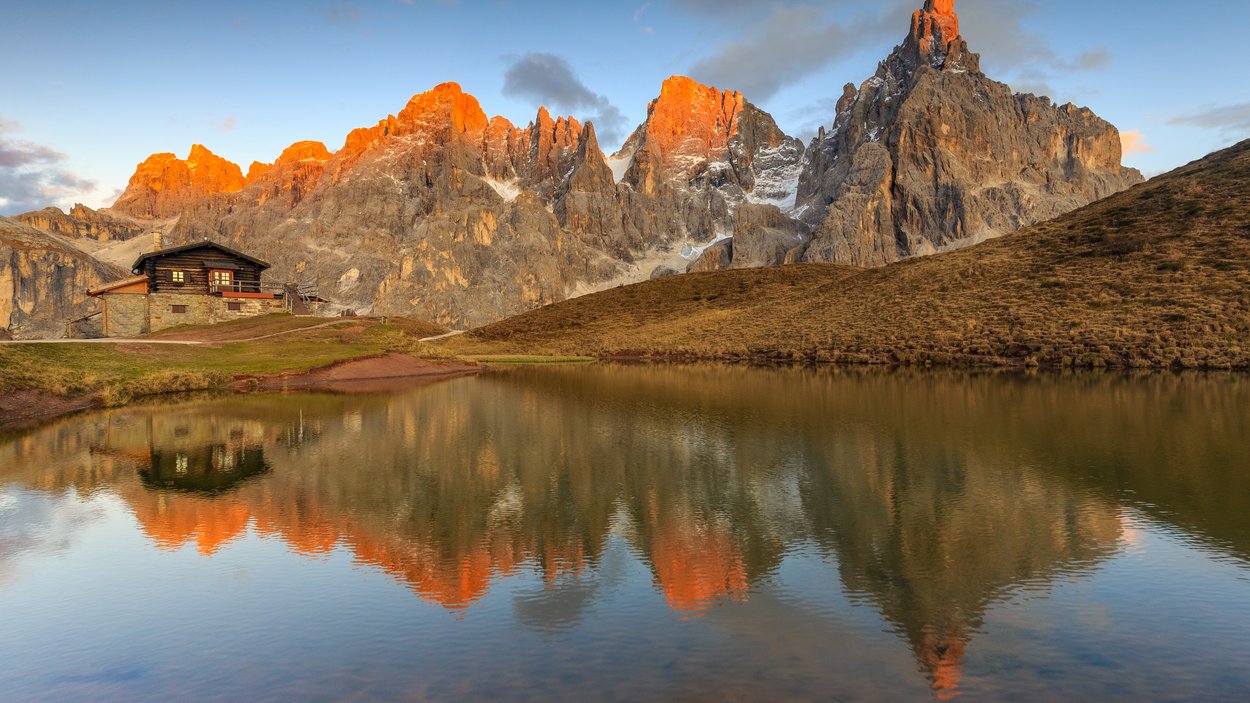Die Alpenpässe in und um Südtirol Almhütte mit Bergspitzen im warmen Abendlicht und Spiegelung im See