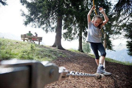 Südtiroler Familienhotels – Lust auf einen Tapetenwechsel? Mädchen balanciert auf einer Seilrutsche im Wald, im Hintergrund sitzen zwei Personen auf einer Bank