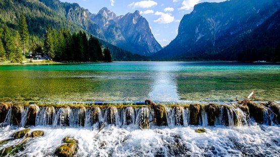In den Hotels in Toblach das Südtiroler Flair genießen Klares Bergseenwasser mit kleinem Wasserfall und bewaldeten Bergen im Hintergrund