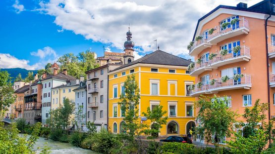 Il vostro hotel a Brunico! Case colorate lungo un fiume in una città sotto un cielo nuvoloso