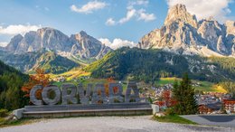 Hotel sulle Dolomiti Scritta in legno Corvara davanti a paesaggio montano e villaggio nelle Dolomiti