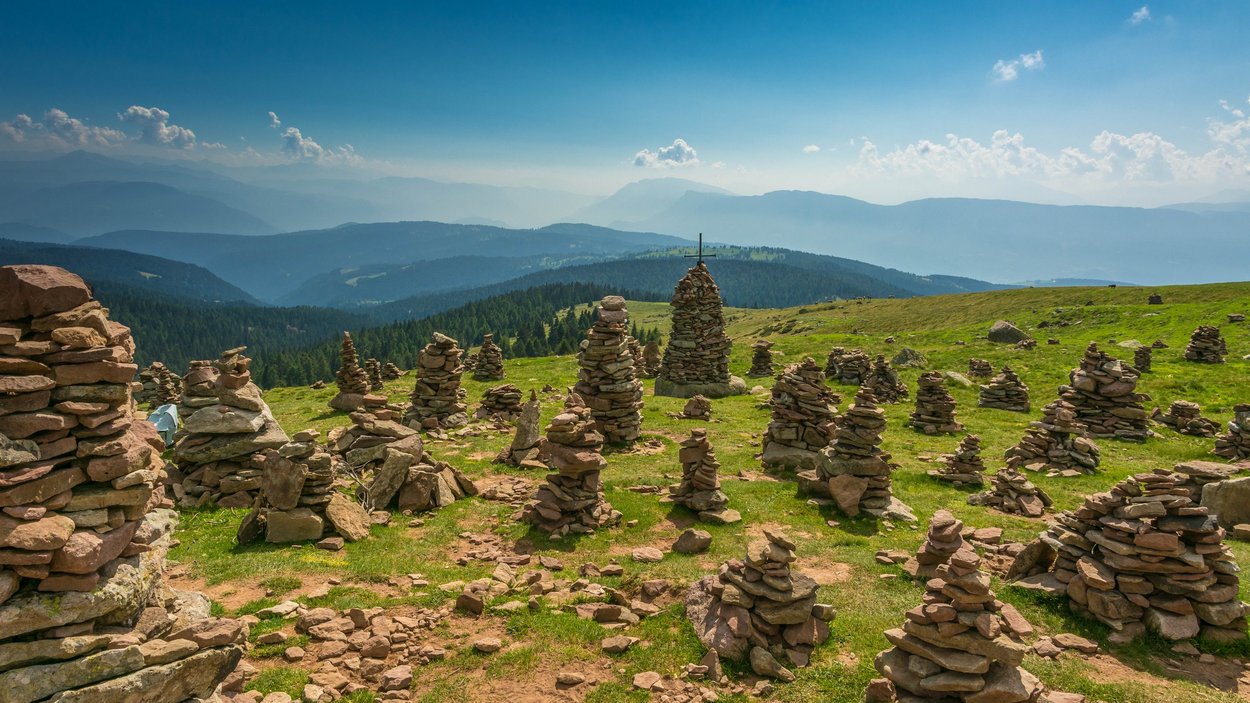 Motorradtouren durch die Alpen – Abenteuer pur Viele Steinmännchen auf einer grünen Bergwiese mit Wald und Bergen im Hintergrund