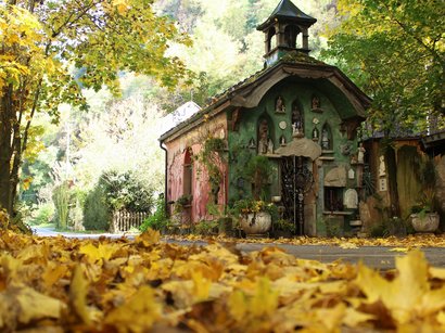 Mondotreno a Rablà Piccola cappella colorata circondata da foglie d'autunno e alberi lungo una strada