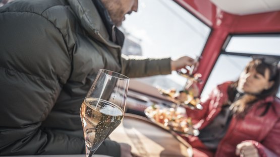 Spaß für die ganze Familie Zwei Personen in Winterjacken mit Sektglas und Snacks in geschlossener Seilbahn