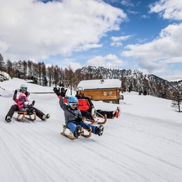 Drei Zinnen-Schneeschuhwanderung Familie rodelnd im Schnee vor einer Berghütte und bewaldeten Bergen