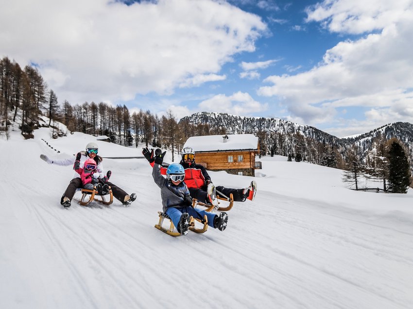 Hotel in Alto Adige 😎☀️ Per vacanze indimenticabili Famiglia che slitta sulla neve davanti a una baita e montagne boschive