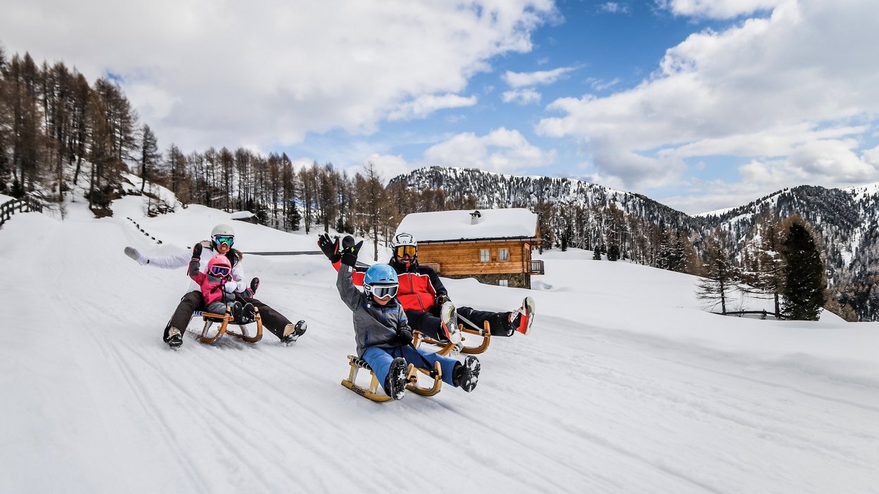 Slittino in Alto Adige: come far battere forte il cuore. Famiglia che slitta sulla neve davanti a una baita e montagne boschive