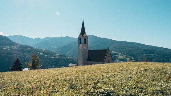 Die Hotels in Feldthurns stehen für einen traumhaften Urlaub Kirchturm auf einer grünen Wiese vor Bergen bei sonnigem Himmel