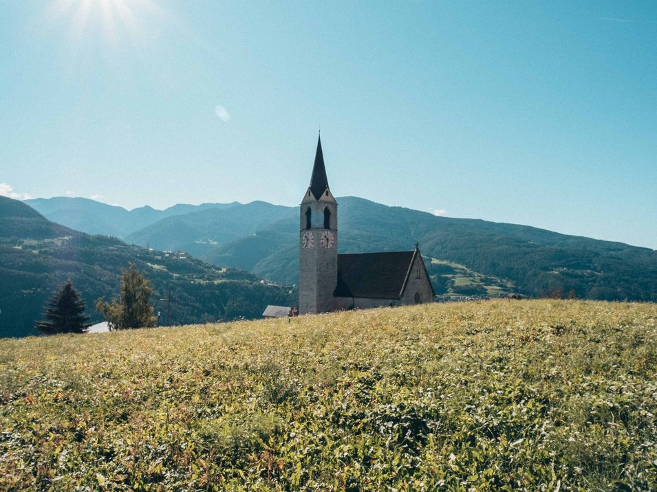 Die Hotels in Feldthurns stehen für einen traumhaften Urlaub Kirchturm auf einer grünen Wiese vor Bergen bei sonnigem Himmel