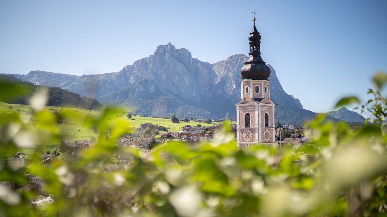 Hotel a Castelrotto sull’Alpe di Siusi Campanile con orologio davanti a paesaggio montano e prati verdi in Alto Adige