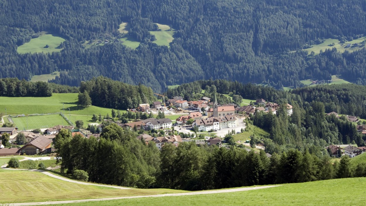 Hotels Terenten: Urlaub im Sonnendorf Bergdorf mit Kirche und grünen Wiesen vor bewaldetem Gebirgszug