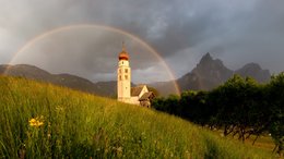 Hotel sulle Dolomiti Chiesa su collina verde con arcobaleno e montagne sullo sfondo