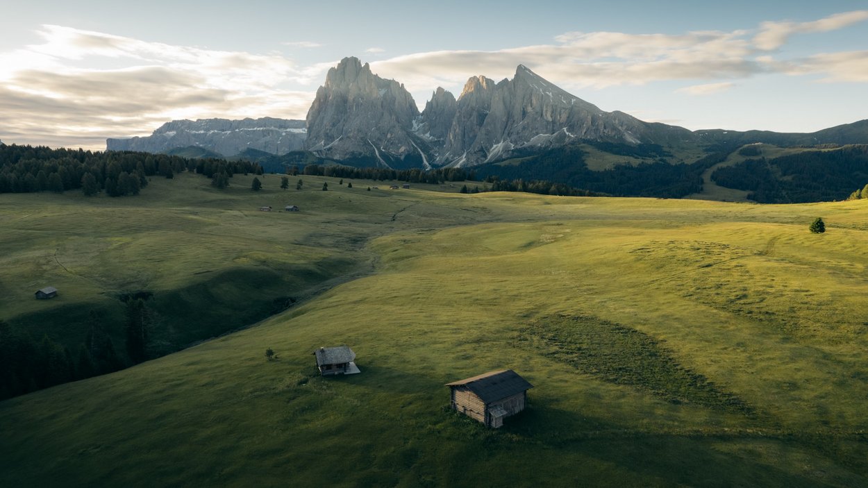 Sole, montagna, Alpe di Siusi! Prato verde con piccole baite davanti a montagne rocciose sotto cielo nuvoloso