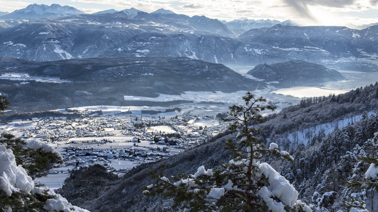 Hotel a Bolzano e dintorni Valle innevato con villaggio, montagne e lago sullo sfondo con cielo nuvoloso
