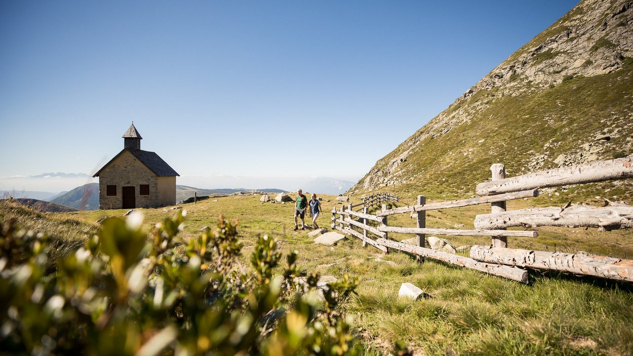 Sporthotels in Südtirol - Aktiv & Entspannt Zwei Wanderer neben Holzzaun in alpiner Landschaft mit Kapelle im Hintergrund