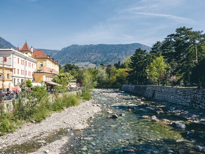 Hotels in Meran und Umgebung Fluss mit klaren Steinen, Baumreihe und historischen Gebäuden unter blauem Himmel