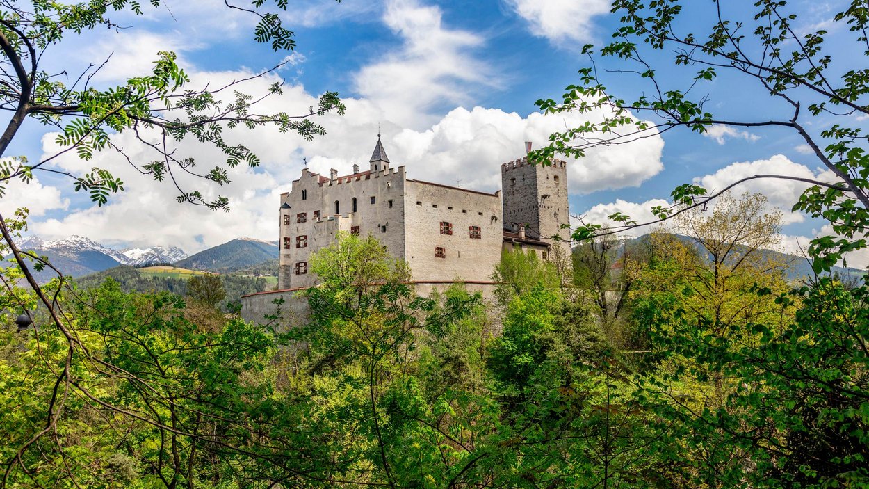Ihre Hotels in Bruneck finden Sie hier! Historisches Schloss umgeben von grünen Bäumen und Bergen im Hintergrund