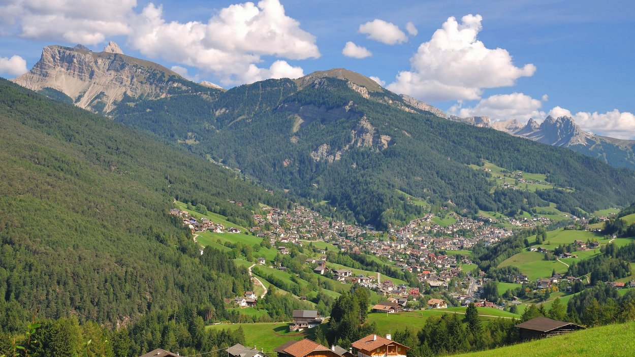 Hotel a Ortisei in Val Gardena Villaggio di montagna con case e prati verdi davanti a montagne boschive e cielo azzurro