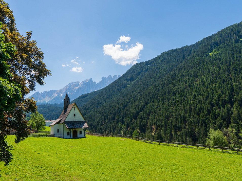Hotels in Welschnofen an der Dolomitenstraße Kleine Kirche auf grüner Wiese vor bewaldetem Berg und blauem Himmel