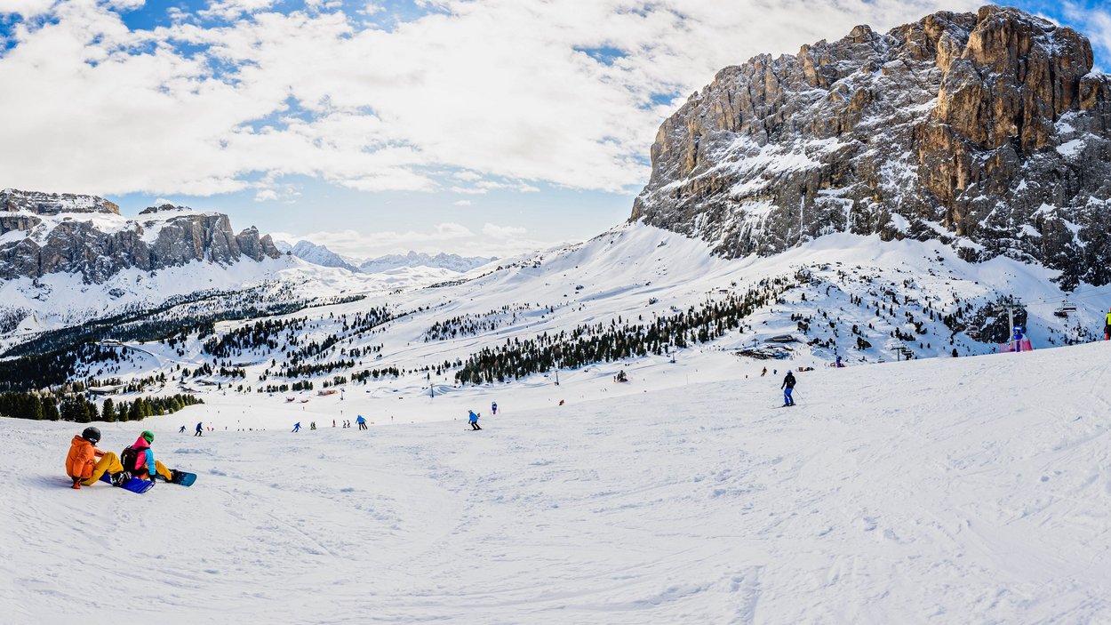 Einmal rund ums Sellamassiv: die Sellaronda Skifahrer und Snowboarder auf verschneiter Bergpiste mit Felsen und Wolkenhimmel