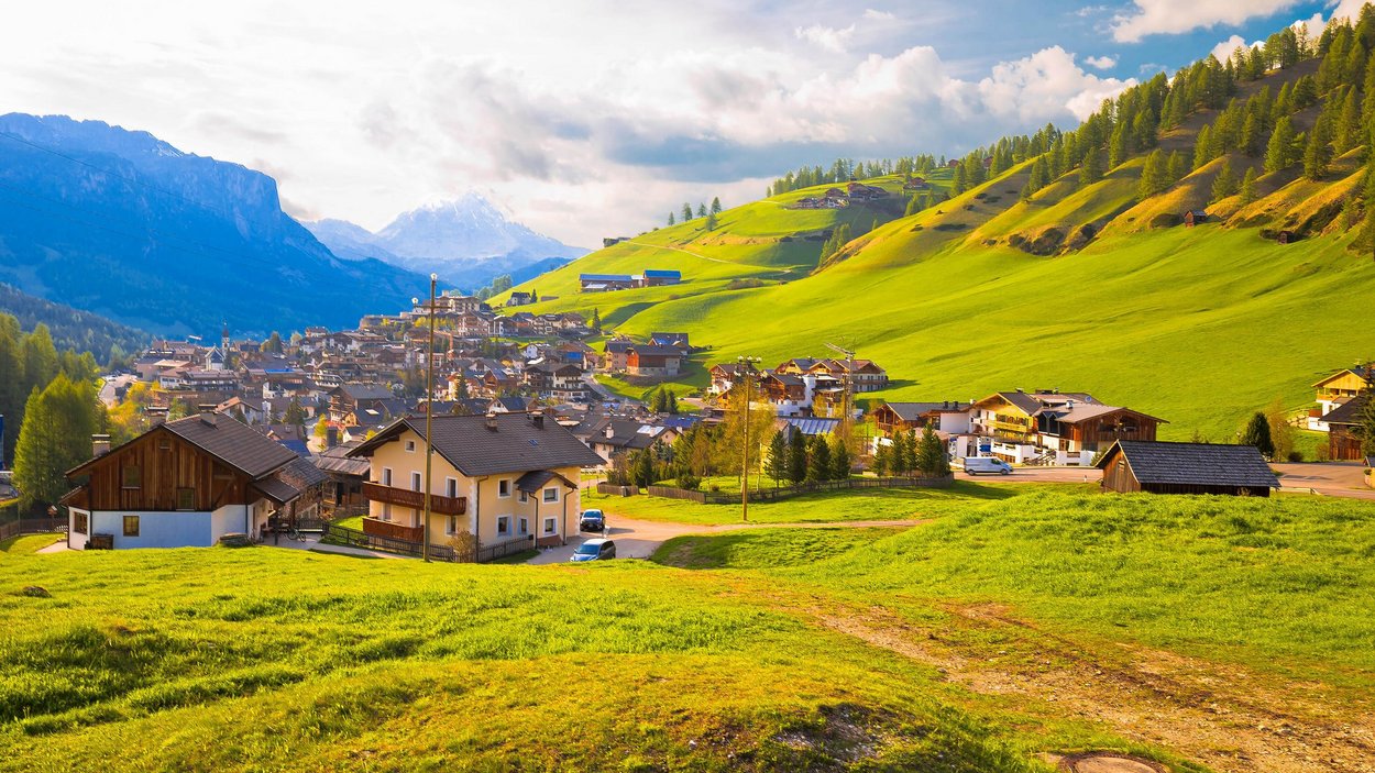 Die Hotels in St. Kassian machen Wanderer glücklich Dorf in grünen Hügeln mit Bergen im Hintergrund bei sonnigem Himmel