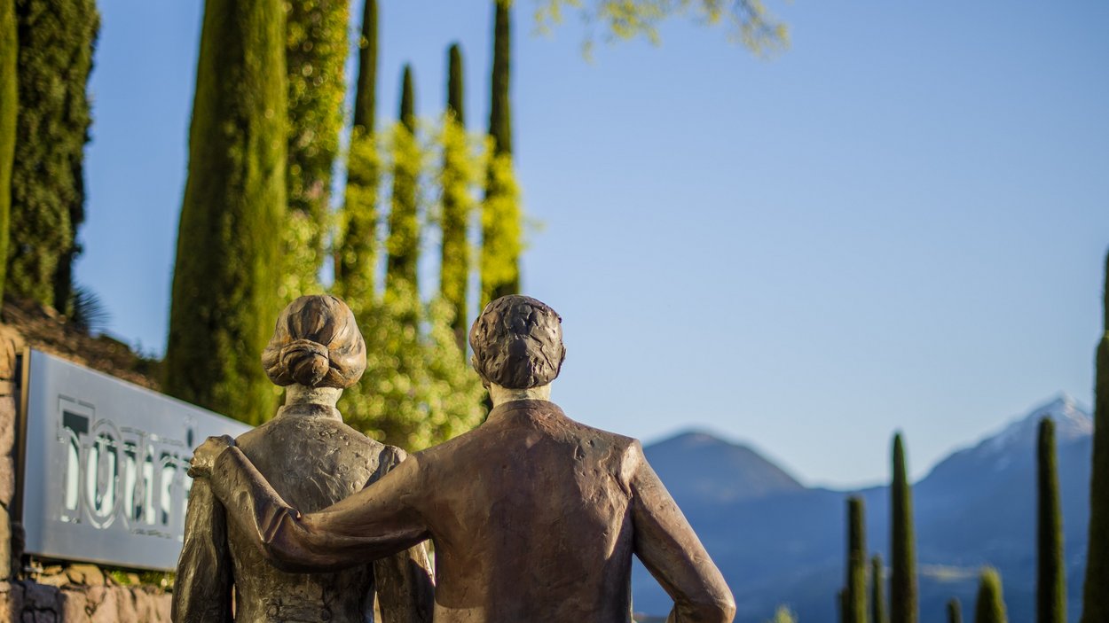 I Giardini di Castel Trauttmansdorff Scultura in bronzo di due persone con il braccio intorno alla spalla davanti a cipressi e montagne