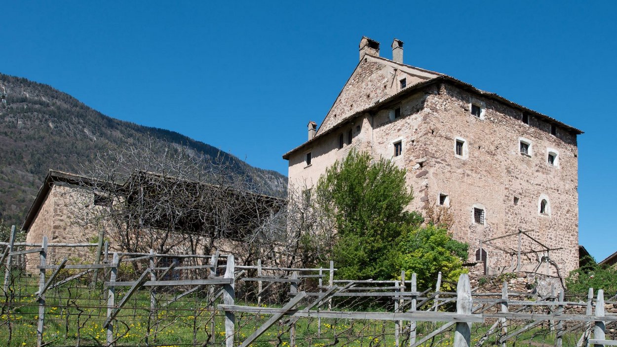 Castel Palù-Schulthaus ad Appiano Vecchia casa di pietra davanti a una montagna alberata sotto un cielo blu limpido