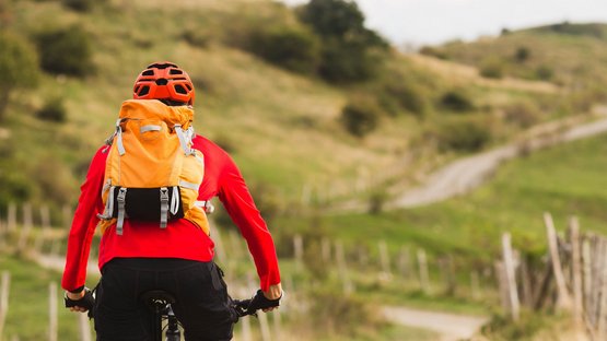 Hotels in Pfalzen: Urlaub an der Pustertaler Sonnenstraße Radfahrer mit orangefarbenem Rucksack fährt auf einem Landweg in hügeliger Landschaft