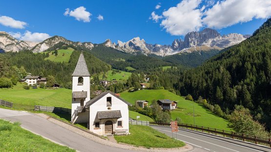 Suchen Sie ein Hotel in Tiers am Südtiroler Rosengarten? Kleine Kirche an einer Straße in grüner alpiner Landschaft mit Bergen im Hintergrund