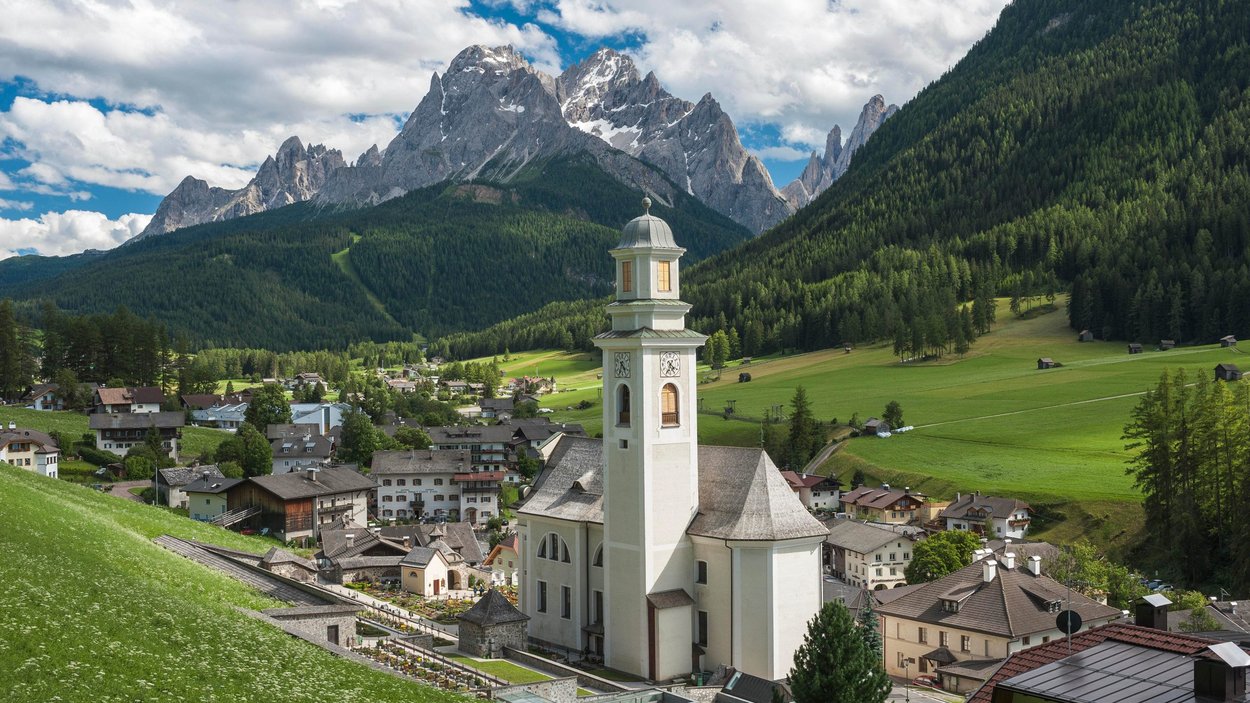 Ihre Hotels in Sexten: traumhafter Urlaub garantiert! Blick auf ein Dorf mit Kirche vor bewaldeten Bergen und Alpen im Hintergrund
