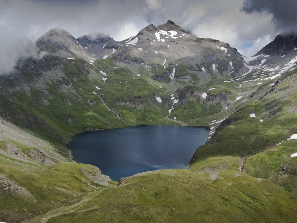 Ein Berg, zwei Gipfel Bergsee umgeben von grünen Bergen und Schneeflecken unter bewölktem Himmel