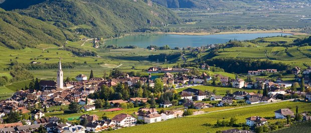 Hotel in Alto Adige 😎☀️ Per vacanze indimenticabili Vista di un paese con chiesa, vigneti e un lago tra le montagne