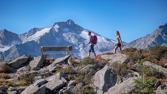 Ihre traumhaften Hotels im Ahrntal in Südtirol Zwei Wanderer gehen auf felsigem Bergpfad mit Bank und Alpenim Hintergrund