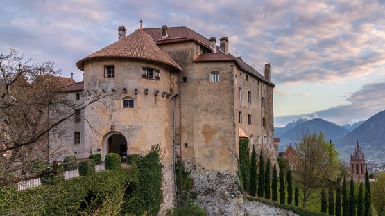 Die schönsten Schlösser in Südtirol: Schloss Schenna Altes Schloss auf einem Hügel mit Blick auf Tal und Berge bei bewölktem Himmel