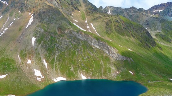 Eisbruggsee – Gletschersee in den Pfunderer Bergen Bergsee mit etwas Schnee an grünen Berghängen unter bewölktem Himmel