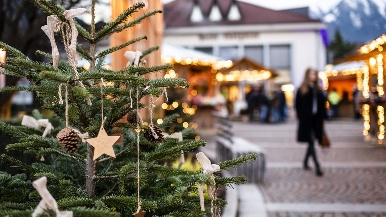 Sarner Alpenadvent – Weihnachten in Südtirol Weihnachtsbaum mit Holzstern und Tannenzapfen vor festlich beleuchtetem Weihnachtsmarkt