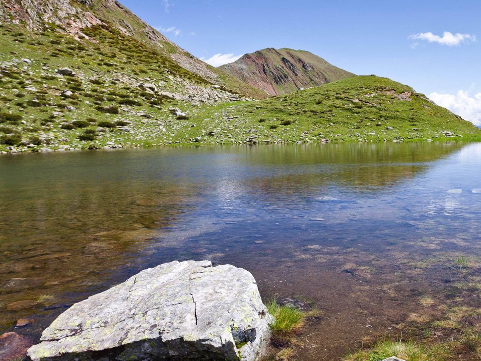 Tra la Val Venosta e la Val d’Ultimo Escursionista accanto a un lago di montagna con erba verde e cielo azzurro