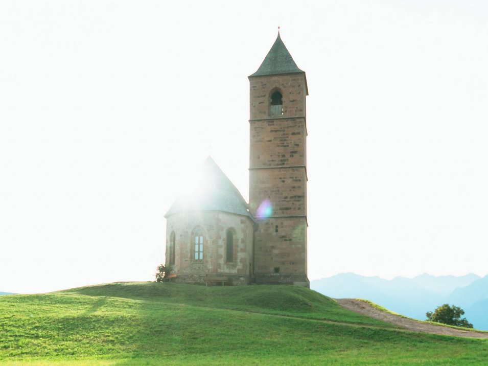Hotels in Hafling: idyllisch, echt, Südtirol Kirche auf grünem Hügel mit Sonnenlicht im Hintergrund