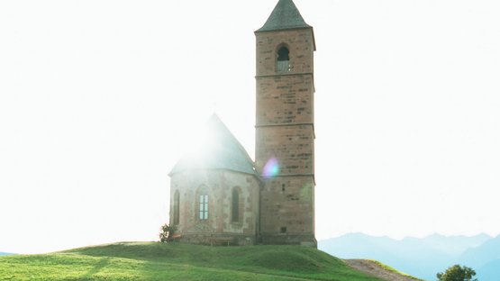 Hotels in Hafling: idyllisch, echt, Südtirol Kirche auf grünem Hügel mit Sonnenlicht im Hintergrund