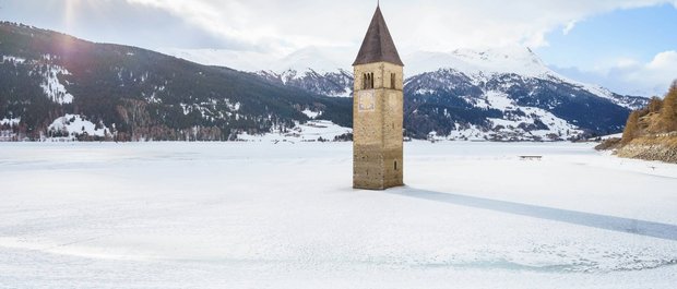 Geheimtipp Hotels in Südtirol 😎☀️ Kirchturm ragt aus zugefrorenem See mit schneebedeckten Bergen im Hintergrund