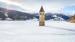 Hotels in Alta Badia: Urlaub durch und durch Kirchturm ragt aus zugefrorenem See mit schneebedeckten Bergen im Hintergrund