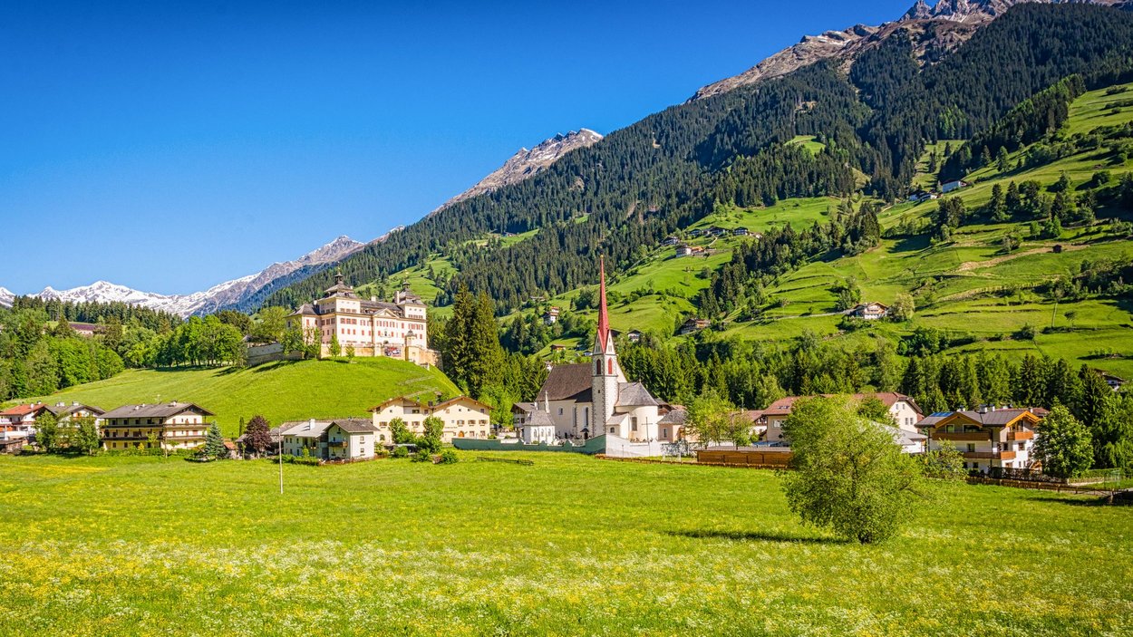 Hotel a Ridanna in Alto Adige Villaggio montano con chiesa e castello davanti a colline verdi e montagne innevate