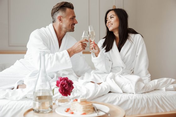 Adults-only-Hotels in Südtirol Paar in Bademänteln stoßt auf dem Bett mit Sektgläsern an